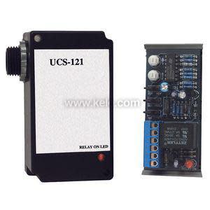 Rectangular black UCS‑121 sensor module with white label and red LED, shown alongside exposed circuit board with blue terminal blocks.